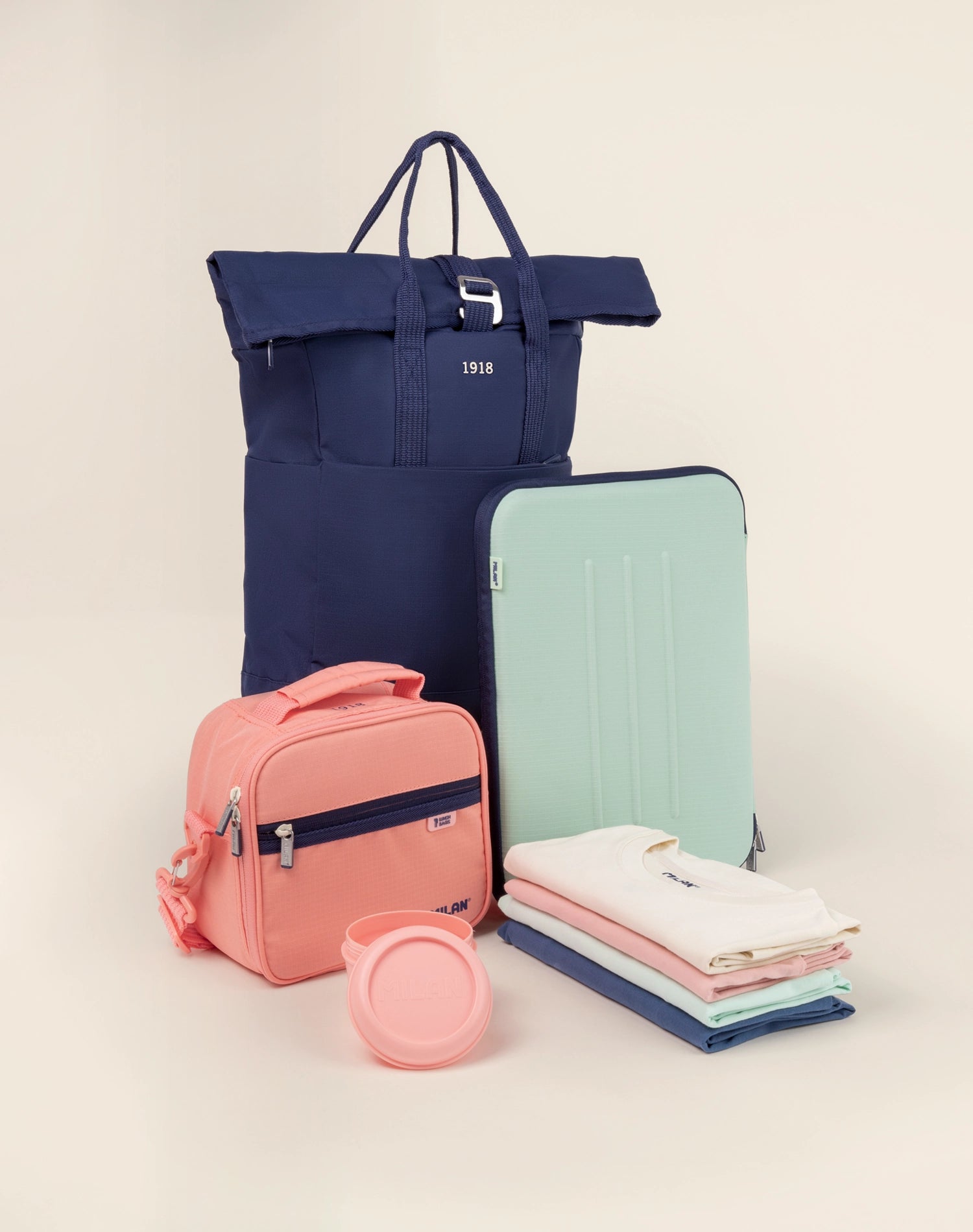 Bodegón de la colección 1918 de MILAN que muestra una variedad de productos, incluyendo bolsas porta alimentos, camisetas de varios colores, una mochila enrollable urbana y una funda para portátil